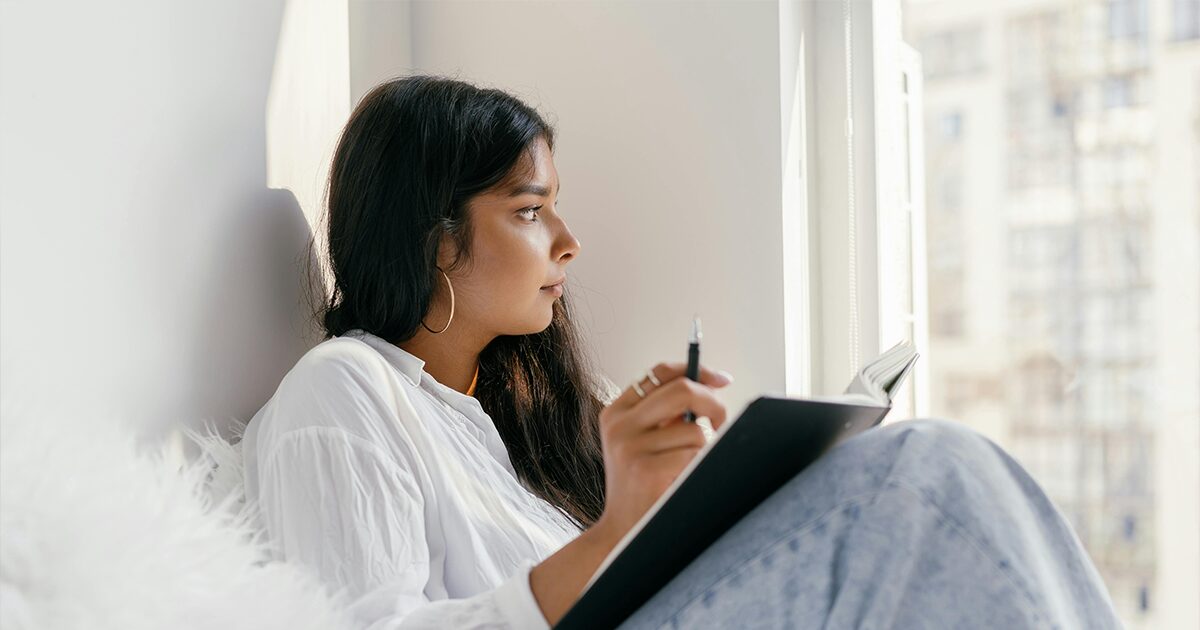 Woman sitting by a window holding a notebook and pen while thoughtfully researching the differences between medical and surgical abortion before making a pregnancy decision.